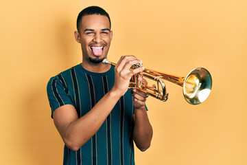 Young african american man playing trumpet sticking tongue out happy with funny expression.