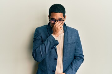 Young handsome hispanic man wearing elegant clothes and glasses smelling something stinky and disgusting, intolerable smell, holding breath with fingers on nose. bad smell