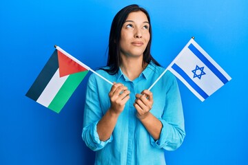 Young latin woman holding palestine and israel flags smiling looking to the side and staring away thinking.