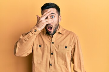 Young hispanic man wearing casual clothes peeking in shock covering face and eyes with hand, looking through fingers afraid