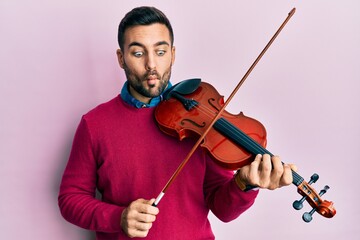 Young hispanic musician man playing violin making fish face with mouth and squinting eyes, crazy...