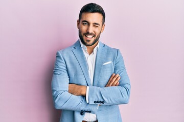 Young hispanic businessman wearing suit with arms crossed gesture winking looking at the camera...