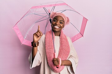 Young african american woman holding umbrella surprised with an idea or question pointing finger...