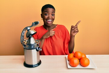 Young african american woman sitting on the table using juicer smiling and looking at the camera pointing with two hands and fingers to the side.