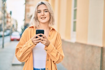 Young blonde girl smiling happy using smartphone at the city.