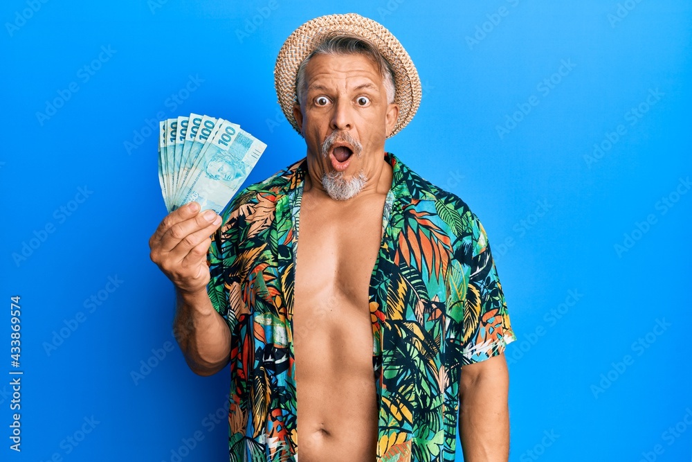 Sticker middle age grey-haired man wearing summer style holding brazilian real banknotes scared and amazed w