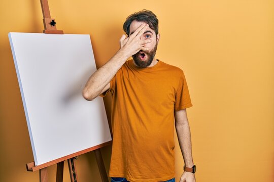 Caucasian man with beard standing by painter easel stand peeking in shock covering face and eyes with hand, looking through fingers afraid