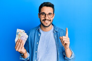 Young hispanic man holding singapore dollars banknotes smiling with an idea or question pointing...