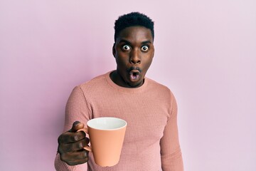 Handsome black man drinking a cup of coffee scared and amazed with open mouth for surprise,...