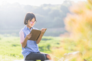 Cute asian little girl with glasses reading a book in garden at summer time