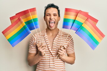 Handsome man wearing make up holding rainbow lgbtq flags sticking tongue out happy with funny expression.
