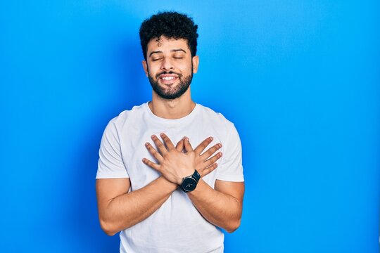 Young arab man with beard wearing casual white t shirt smiling with hands on chest with closed eyes and grateful gesture on face. health concept.