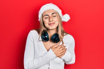 Beautiful hispanic woman wearing christmas hat and headphones smiling with hands on chest, eyes closed with grateful gesture on face. health concept.