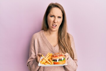 Young blonde woman eating a tasty classic burger with fries in shock face, looking skeptical and sarcastic, surprised with open mouth