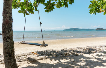 wooden swing hanging on the tree on brow sand beach, blue sea under blue sky on background