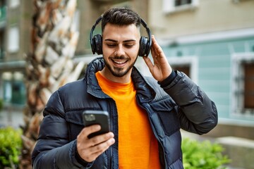 Handsome hispanic man with beard smiling happy and confident at the city wearing winter coat listening to music from smartphone