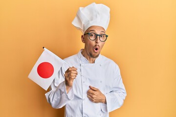 Bald man with beard wearing professional cook uniform holding japan flag scared and amazed with open mouth for surprise, disbelief face