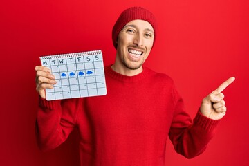 Bald man with beard holding rainy weather calendar smiling happy pointing with hand and finger to the side