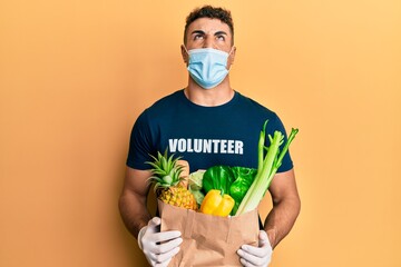 Hispanic young man wearing medical mask holding paper bag with groceries angry and mad screaming frustrated and furious, shouting with anger looking up.