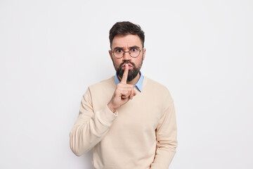 Serious bearded man makes silence gesture prohibits to speak shows hush sign has strict face expression wears casual clothes isolated over white background asksto keep voice down scolds bad behavior - obrazy, fototapety, plakaty