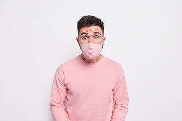 Personal protection and quarantine concept. Startled young man wears face mask to protect from coronavirus hears shocking news dressed in jumper isolated over white background. Viral infection