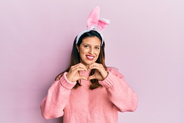 Beautiful hispanic woman wearing cute easter bunny ears smiling in love doing heart symbol shape with hands. romantic concept.