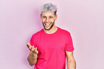 Young hispanic man with modern dyed hair wearing casual pink t shirt smiling cheerful with open arms as friendly welcome, positive and confident greetings - obrazy, fototapety, plakaty