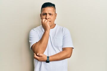 Young latin man wearing casual white t shirt looking stressed and nervous with hands on mouth biting nails. anxiety problem.