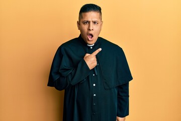 Young latin priest man standing over yellow background surprised pointing with finger to the side, open mouth amazed expression.