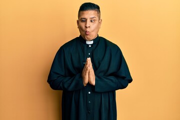 Young latin priest man praying with hands together making fish face with mouth and squinting eyes,...