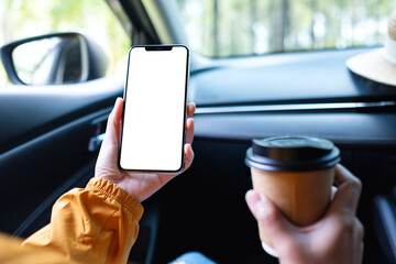 Mockup image of a woman holding and using mobile phone with blank screen while drinking coffee in the car