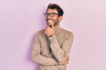 Handsome man with beard wearing turtleneck sweater and glasses with hand on chin thinking about...