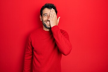 Handsome man with beard wearing casual red sweater covering one eye with hand, confident smile on...