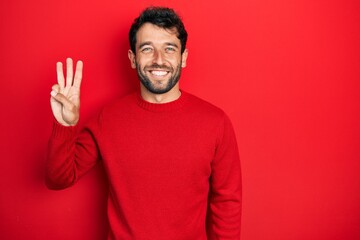 Handsome man with beard wearing casual red sweater showing and pointing up with fingers number three while smiling confident and happy.