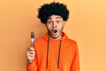 Young african american man with afro hair holding one silver fork scared and amazed with open mouth...