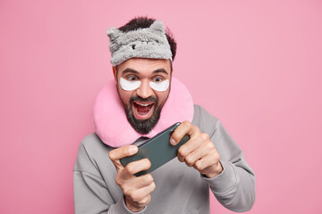 Cheerful bearded man plays video games on smartphone before going to bed wears sleepmask and comfortable pillow around neck beauty patches addicted to modern technologies tries to pass hard level.