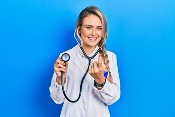 Beautiful young blonde doctor woman holding stethoscope beckoning come here gesture with hand...