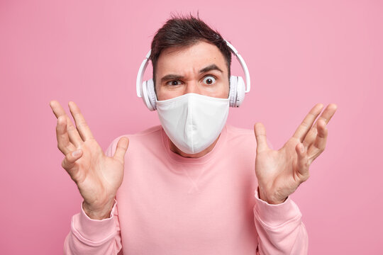 Puzzled confused man raises palms listens music via white wireless headphones raises eyebrows wears protective face mask to prevent coronavirus dressed casually isolated over pink background