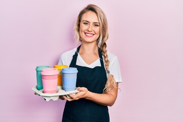 Beautiful young blonde woman wearing waitress apron holding take away cup of coffees smiling with a...