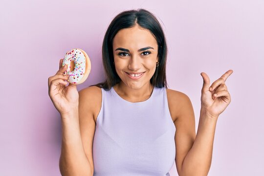 Young brunette woman holding tasty colorful doughnut smiling happy pointing with hand and finger to the side