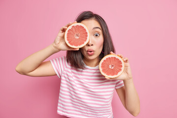 Surprised young Asian woman poses with citrus fruit indoor holds two halves of fresh grapefruit...