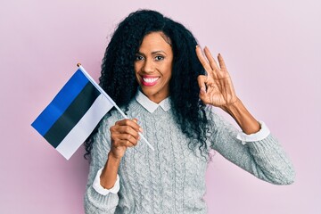 Middle age african american woman holding estonia flag doing ok sign with fingers, smiling friendly gesturing excellent symbol