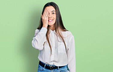 Young brunette teenager wearing business white shirt covering one eye with hand, confident smile on face and surprise emotion.