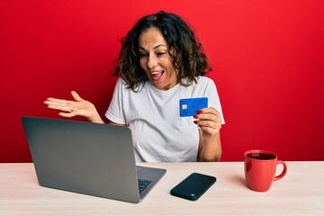 Beautiful middle age woman working at the office using laptop and credit card celebrating achievement with happy smile and winner expression with raised hand