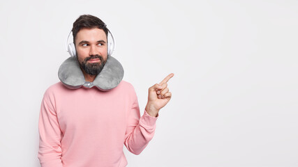 Handsome pleased bearded man points away on blank space demonstrates something grabs your attention to advertisement wears neck pillow for comfortable rest listens music via stereo headphones