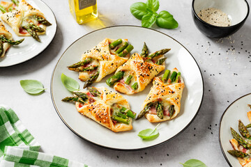 Baked green asparagus with ham and cheese in puff pastry sprinkled with sesame seeds and green basil leaves. Light gray stone background.