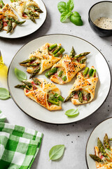 Baked green asparagus with ham and cheese in puff pastry sprinkled with sesame seeds and green basil leaves. Light gray stone background.