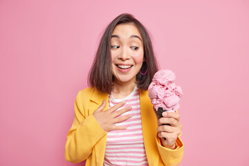 Photo of good looking young Asian woman with dark hair holds yummy ice cream in waffle cone smiles gladfully dressed in stylish outfit wants to eat delicious frozen summer dessert. Enjoy it.