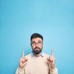 Serious bearded brunet adult man focused above indicated with index fingers overhead shows blank...