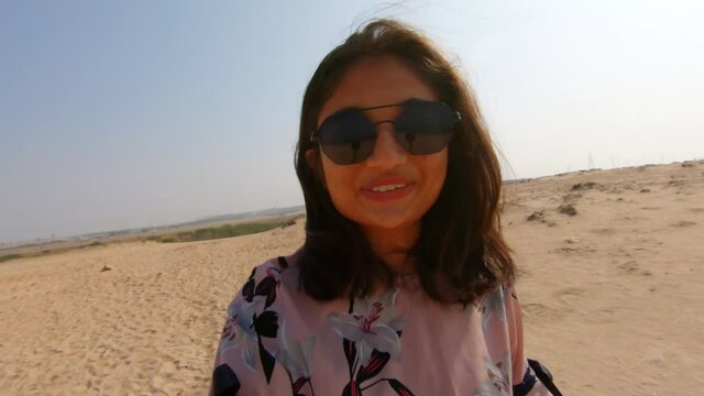 Happy young Indian woman vlogger wearing sunglasses taking a walk at beach and talking while looking at camera. Female travel vlogger at beach in summer